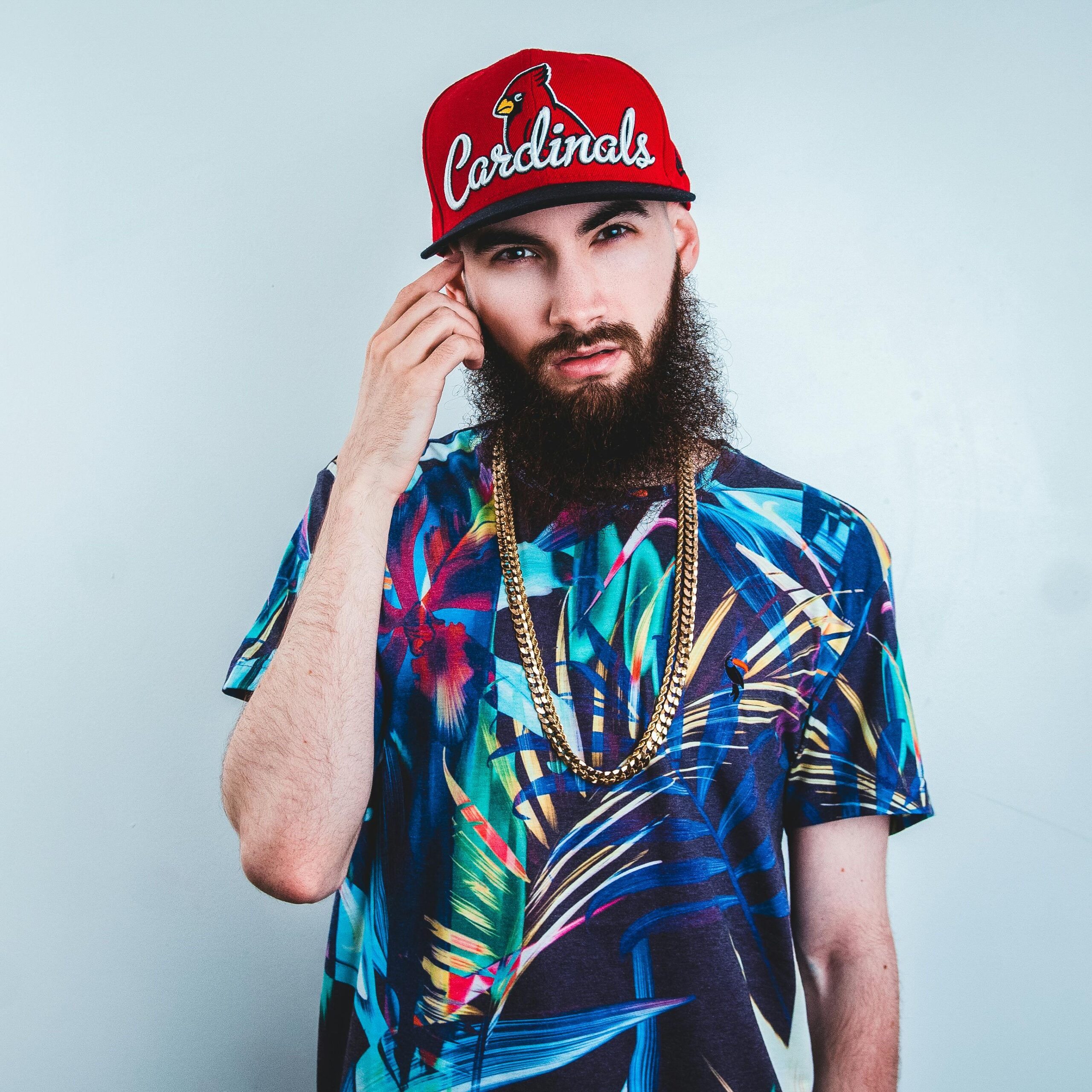 Homme barbu avec casquette rouge et t-shirt coloré, chaîne dorée autour du cou.